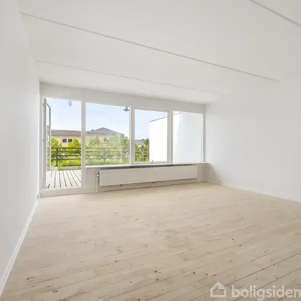En tom stue med lyse trægulve og hvide vægge, med store vinduer og en åben dør ud til en balkon, der giver udsigt til en grøn have.