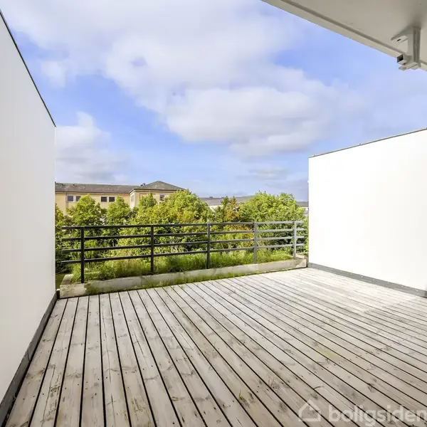En rummelig terrasse med trægulv strækker sig ud mod en grøn udsigt. Den er omgivet af hvide vægge, og en sort gelænder beskytter kanten. Skyer og blå himmel ses ovenfor.