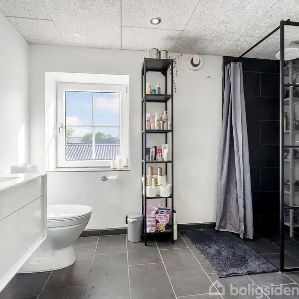 Et moderne badeværelse med grå fliser, en hvid vask, et toilet, og en brusekabine med glasvægge. Ved siden af vinduet står en sort reol fyldt med toiletartikler.