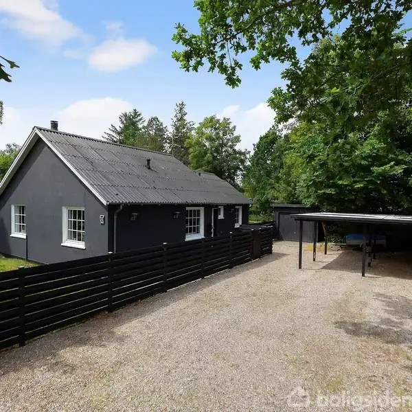 En sortmalet bungalow med hvide vinduer står ved siden af en grusvej. Der er en sort træhegn og en overdækket carport ved siden af huset, omgivet af grønne træer.