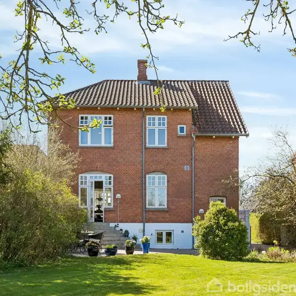 Rødt murstenshus står stille omgivet af en grøn have med træer og buske. Trappen foran huset fører til en åben terrasse. Sollys oplyser scenen under en blå himmel.