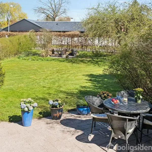 En terrasset med et rundt bord og stole er omgivet af blomsterkrukker foran en grøn græsplæne. Baggrunden har en hæk og træer, med huse i baggrunden.