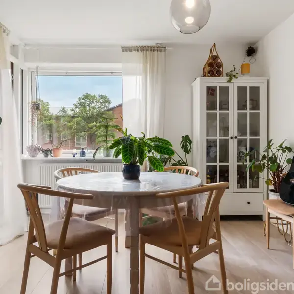 Rundt spisebord står i en lys spisestue med fire stole. Planter pynter rummets vindueskarm og reoler. En akustisk guitar står op ad et skab, og en lampe hænger i loftet.