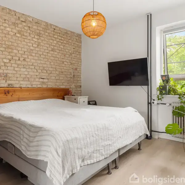 En seng med hvidt sengetøj står i et soveværelse med en rå murstensvæg. Ved siden af er et vindue med grønne planter og en fladskærm på væggen.