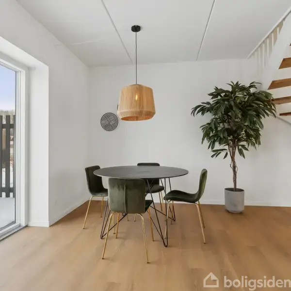 Rundt bord står med tre stole i spisestue med trægulv, stor plante og trappe. To store vinduer til venstre slipper lys ind. Lampe hænger over bordet.