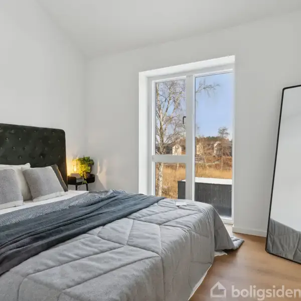 En seng med mørkt sengegavl står i et lyst soveværelse. Sengen har gråt sengetøj. Ved siden af står et spejl op ad væggen og en lampe på et natbord. Udsigt gennem vindue.