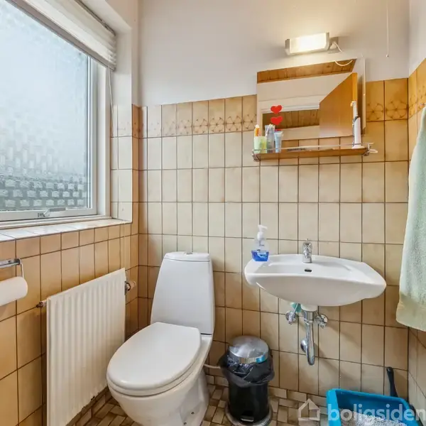 Toilet med hvidt sæde står ved siden af en håndvask med spejl. Væggene er flisebelagte med beige fliser. Et håndklæde hænger på væggen. Rummet har et vindue.