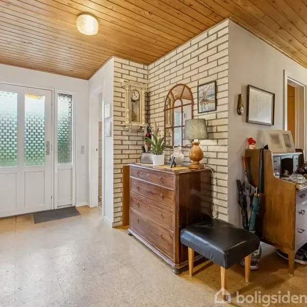 En entré med en kommode, hvorpå der står en lampe og dekorationer. Rummet har træloft og murstensvæg, og der er flere døre og et vindue med glasruder, samt en plante på gulvet.
