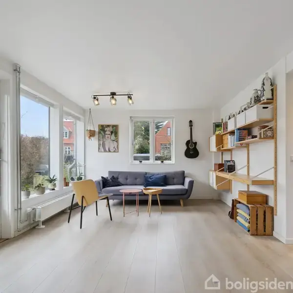 Stue med sofa og stole står ved store, lyse vinduer. Væggen har en guitar og hylder. Rummet er lyst og moderne med udsigt til en have.