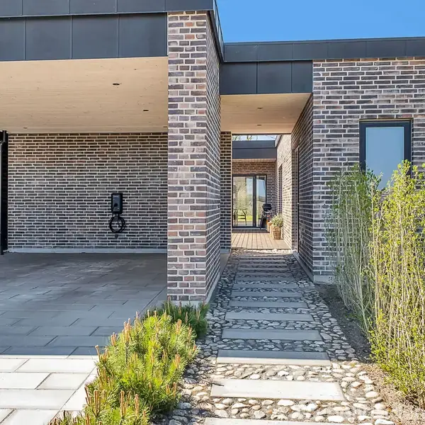 Moderne hus i grå mursten med en carport til venstre. En brolagt sti fører gennem en smal passage til hovedindgangen. Omgivelserne har buske og fliser i en have.