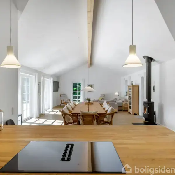 Køkkenø med komfur i forgrunden. Spisebord og stole i moderne, lys stue med hvide vægge og loft. Store vinduer og dør tilgår terrasse. Brændeovn i hjørnet.