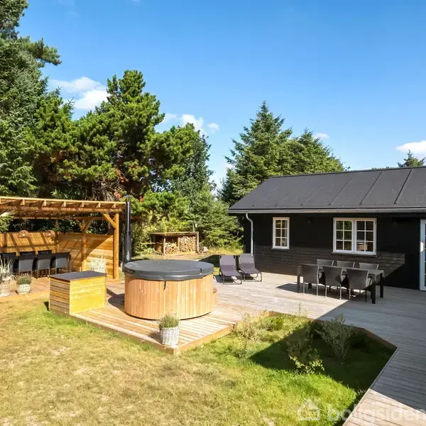 Et sommerhus med sort facade står i en solrig have omringet af høje træer. En træterrasse rummer et bord, stole og et rundt vildmarksbad.