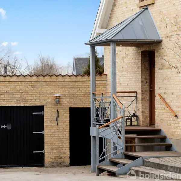 Et gult teglhus har en trappe med gelænder, der fører til en trædør. Garagen ved siden af har sorte døre. Træer og skyer er synlige i baggrunden.