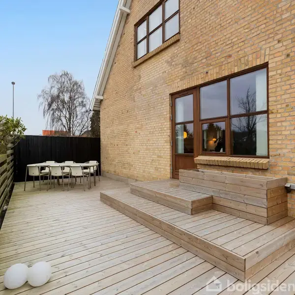 Træterrasse med trin foran et murstenshus. Terrassen har et bord og stole i hjørnet. Træhegn og planter omkranser området på en solrig dag.