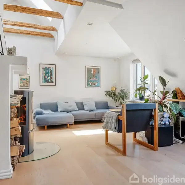 Stue med lyse træmøbler og reol, sofa og billeder på væggen. Store skråvinduer lader lys ind, og planter er placeret langs vindueskarmen. Trægulv og fritlagte træbjælker ses.