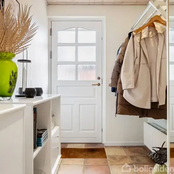 Dør i en gang med mange ophængte frakker og spejl. Grøn vase med tørrede planter på reol med bøger. Flisegulv og lys vægindretning.