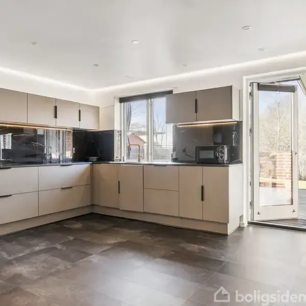 Køkken med beige skabe og sort bordplade, der er u-formet. Der er to vinduer og en terrassedør, der fører ud til en have. Lys, moderne indretning.