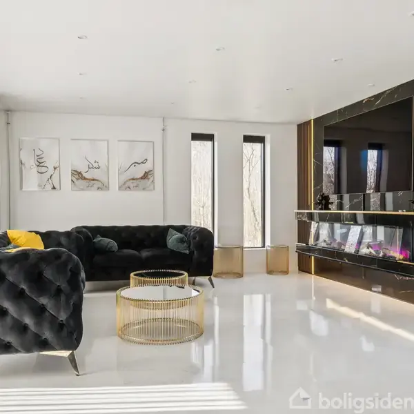 Sorte sofaer og gyldent sofabord står i en moderne stue med hvidt gulv. Væggene har abstrakte malerier, og en stor pejs er centralt placeret under et fladskærms-tv.