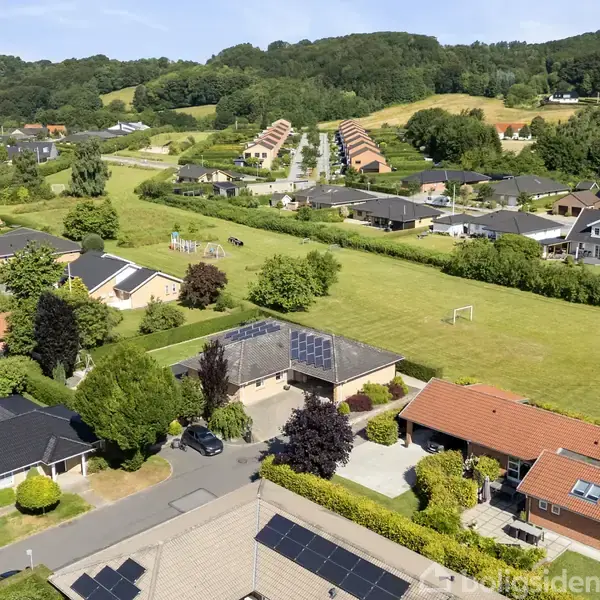 En række huse med røde og sorte tage står langs en vej omgivet af grønne haver. I baggrunden ses en fodboldbane og skovklædte bakker.