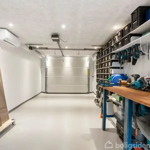 En garage med værktøjsopbevaring er velorganiseret. Langbordet til højre har værktøj og kasser, mens en reol over det er fyldt med bokse. Væggen til venstre har træplanker.