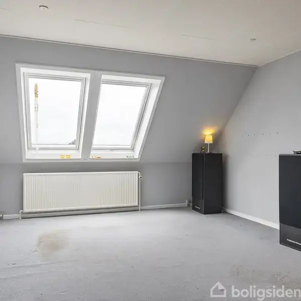 Et tomt rum med lysegrå vægge og gulvtæppe har to skråvinduer. En radiator er under vinduerne. En sort højttaler står til højre med en lampe ovenpå.