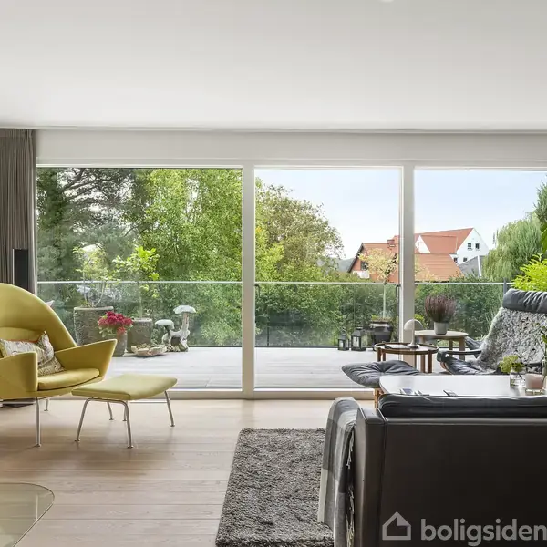 Stue med moderne møblering, herunder en gul stol og sort sofa, står ved siden af store glaspartier. Udsigt til en terrasse og grøn have med rødligt tag i baggrunden.