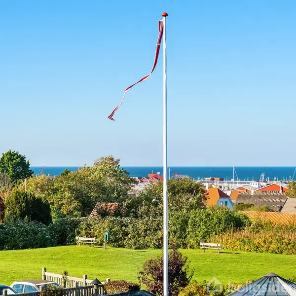 Flagstang med vimpel bøjer i brisen. I baggrunden ses en græsplæne, buske, huse og havet. Himlen er klar og blå.