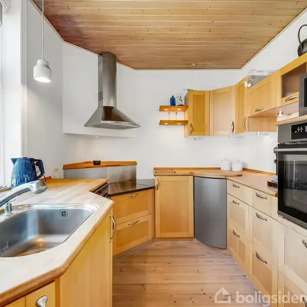 Et L-formet køkken står ubenyttet med træskabe, ovn og mikrobølgeovn indbygget. Lysindfald fra store vinduer oplyser rummet. Et stålvask og emhætte i rustfrit stål kompletterer indretningen.