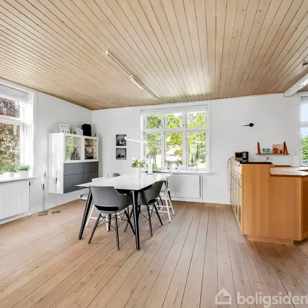 Et lyst køkkenalrum med trægulv og træloft. Et hvidt spisebord og sorte stole står midt i rummet. Store vinduer lader naturligt lys ind, og køkkenet ses til højre.