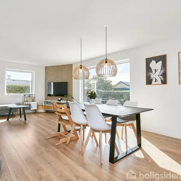 En lys stue med træbord og spisestole. To moderne lamper hænger over bordet. Sofa og fjernsyn i baggrunden. Vægdekorationer og planter skaber en hyggelig atmosfære.