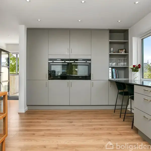 Grå køkkenindretning med indbyggede hvidevarer ovenpå lyse trægulve; siddepladser ved bord. Vinduer åbner til grønne omgivelser. Hylder med glas og bøger ved siden af skabe.
