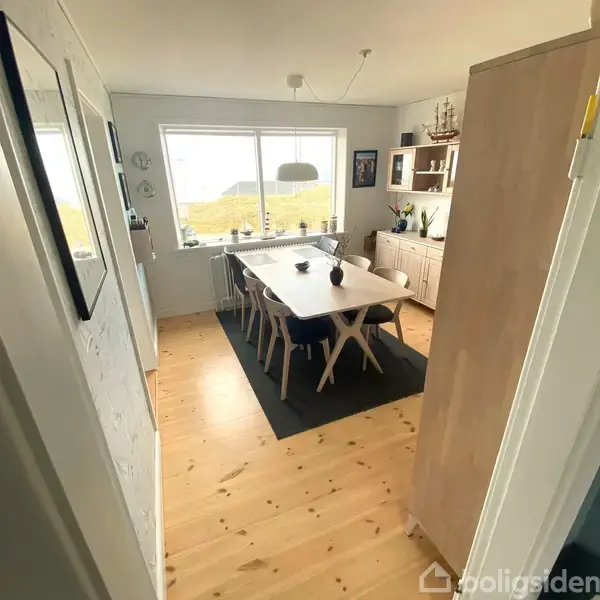 Spisebord med stole står i lys spisestue. Store vinduer giver udsigt til landskab. Rummet er lyst med trægulv og hvid væg, reol og dekorative genstande.