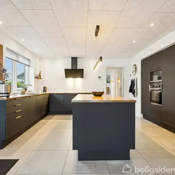 Et moderne køkken med sorte skabe og skuffer, står i et lyst rum med store vinduer. En ø i midten har en træoverflade, omgivet af hvide vægge og lofter.