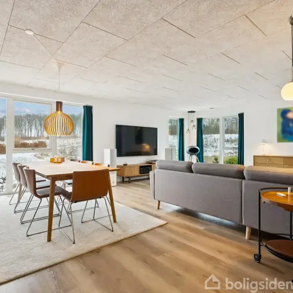 Stuen har en grå sofa og et spisebord med seks stole. Rummet er lyst med store vinduer, der viser en sneklædt have. Loftslamper og træmøbler fuldender indretningen.