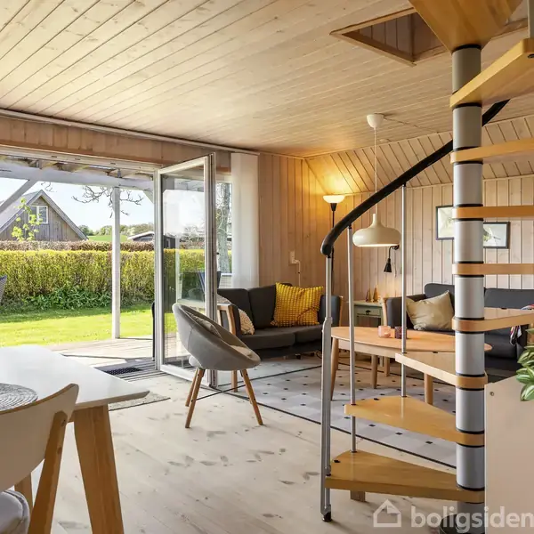 Spiraltrappe i træ fører opad, omgivet af et stueområde med sofagruppe, gulvlampe og spisebord. Store vinduer åbner ud til en grøn have med veltrimmet hæk.