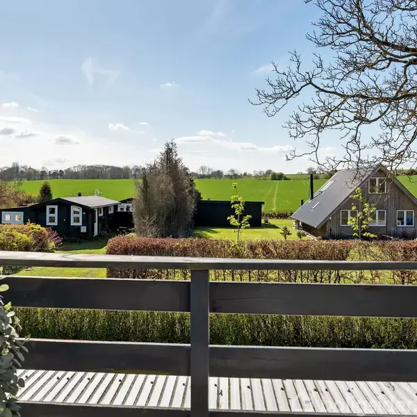 Træterrasse med udsigt over en grøn have og et åbent marklandskab. To huse med skorstene ses i baggrunden under en blå himmel.