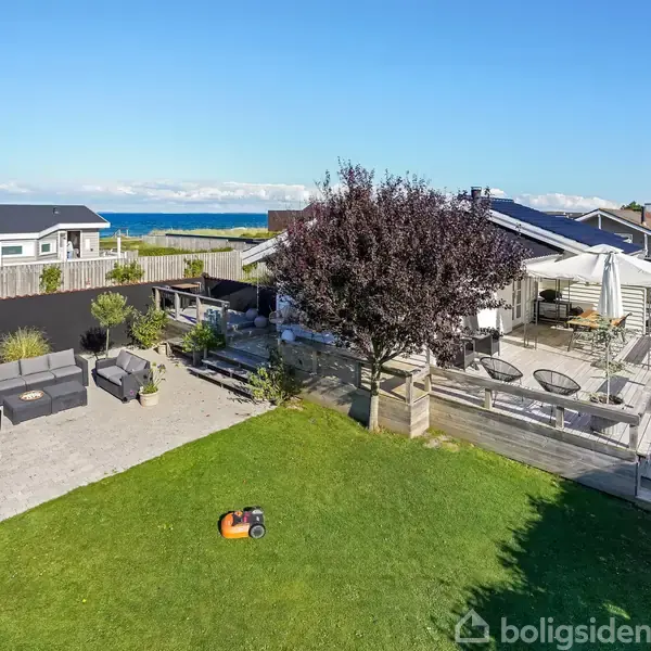 En græsplæne med en robotplæneklipper. I baggrunden ses en terrasse med møbler, et flagstang med dansk flag, og udsigt til havet.