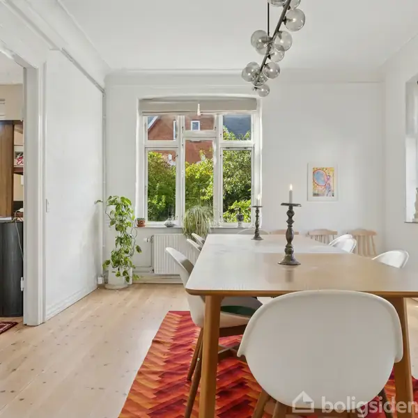 Et spisebord med fire stole står i et lyst rum med trægulve. En lampe hænger over bordet, og vinduer med planter giver udsigt til grønne træer. En guitarkasse står op ad væggen.