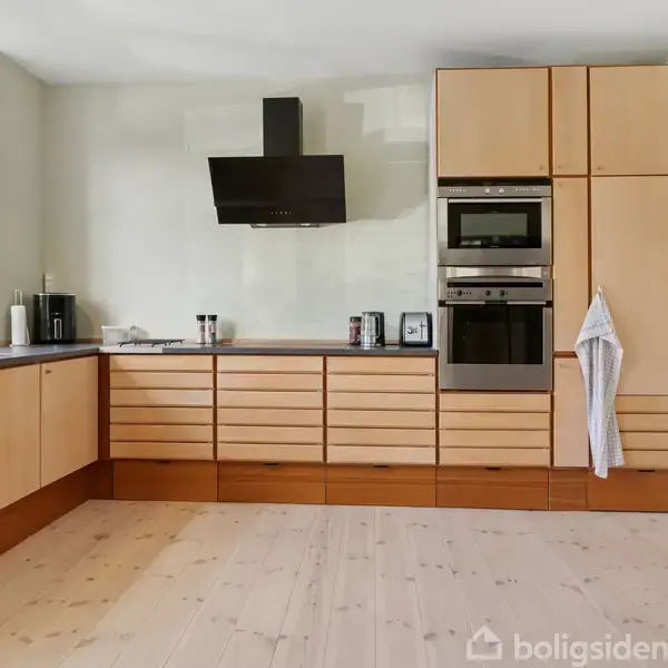 Køkken med lyse træskabe, integreret ovn og mikroovn, og sort emhætte. Krydderurter ved vindue til venstre. Træbordplade og lyst trægulv skaber en lys atmosfære.