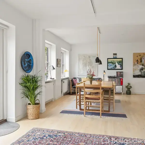 Spisebord med stole står i lys stue med trægulv. Store glasdøre åbner ud til en grøn have. Bøger på reol til højre. Kunstværker hænger på væggene.