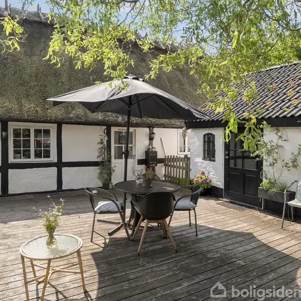 Et bord med stole står på en terrasse under en parasol. Omkring er der et bindingsværkshus med stråtag og en have. Atmosfæren er hyggelig og afslappende.