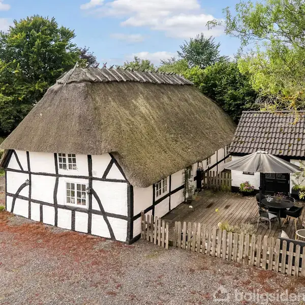 Bindingsværkshus med stråtag omgivet af grusbelagt gårdsplads og træterrasse med møbler og parasol; grønne træer i baggrunden fuldender det landlige, idylliske scenarie.