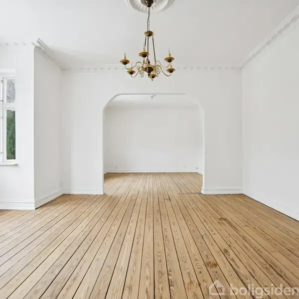 Stort rum med glat trægulv og en gylden lysekrone hængende fra loftet. Rummet er tomt, har hvide vægge og store vinduer, hvilket giver naturligt lys.