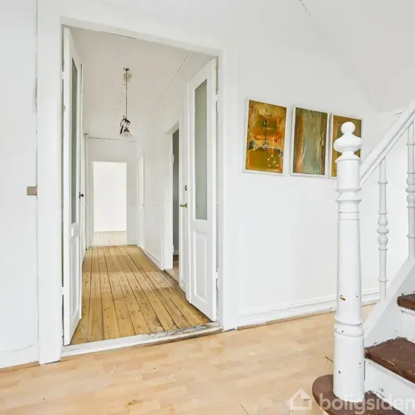 En entré med en trappe til højre, fører til en gang med trægulv og to hængende lamper. To malerier hænger på højre væg. Dør til venstre.