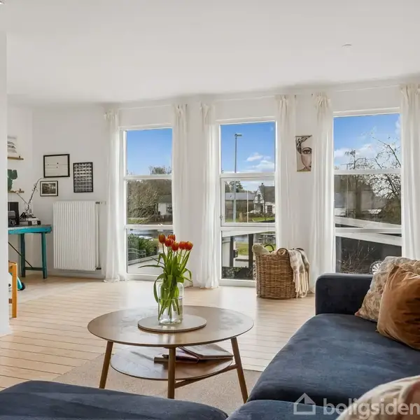 Sofaen med puder står ved et rundt sofabord med blomster i stue med lyse trægulve. Store vinduer med gardiner giver udsigt til en have.