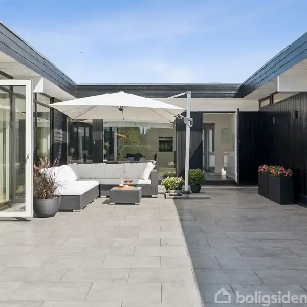 Terrasse med sofaarrangement og parasol står på flisebelagt gård. Omkringliggende moderne bygning med åbne døre og vinduer, som skaber en lys og inviterende atmosfære.