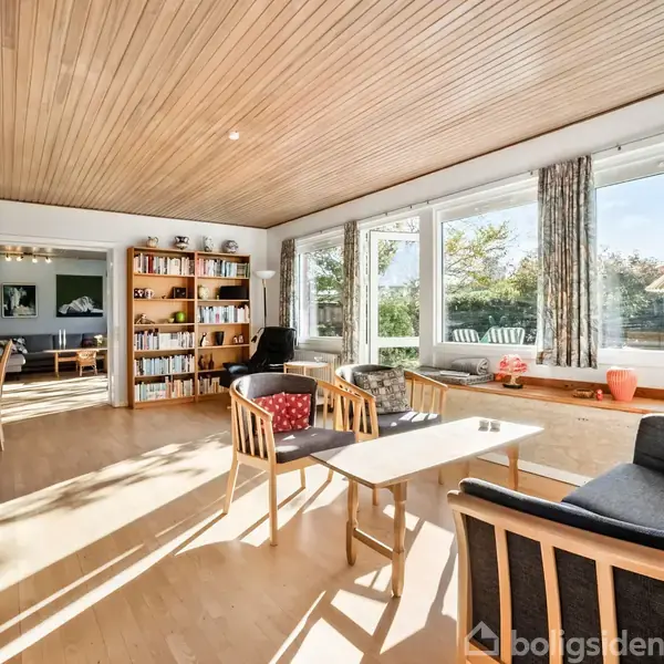 Stue med træmøbler og sofa omkring et bord, beliggende ved store vinduer. Reoler med bøger står mod bagvæggen. Sollys strømmer ind, afslørende en lys og behagelig atmosfære.