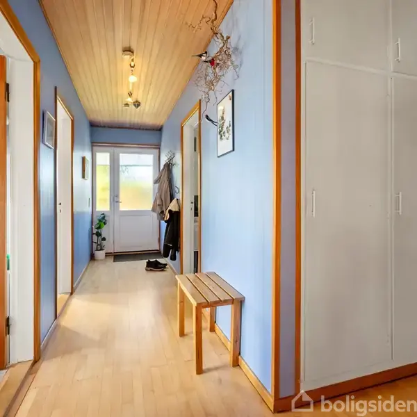 En lang gang med lyseblåt interiør, trægulv og træloft. Døre på begge sider, et lille træbænk til højre, og tøj hængende bagerst. Møbleret med minimalistisk dekoration.