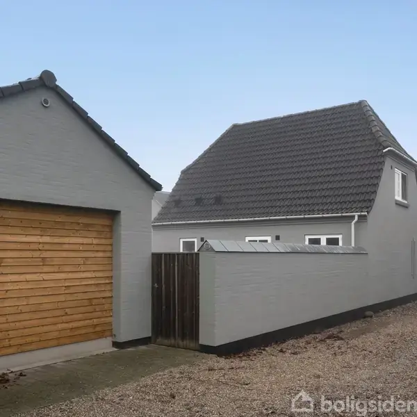 Gråt hus med mørkt tag står ved siden af en garage med træbeklædt port. Omgivet af grusvej og klart, blåligt himmelspanorama.