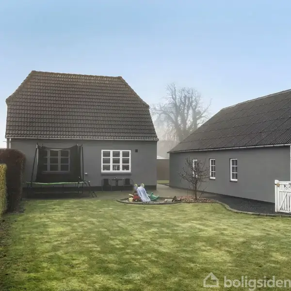 Et gråt hus med rødt tag står i en frodig have. En trampolin er til venstre, og der er en græsplæne omgivet af hæk.